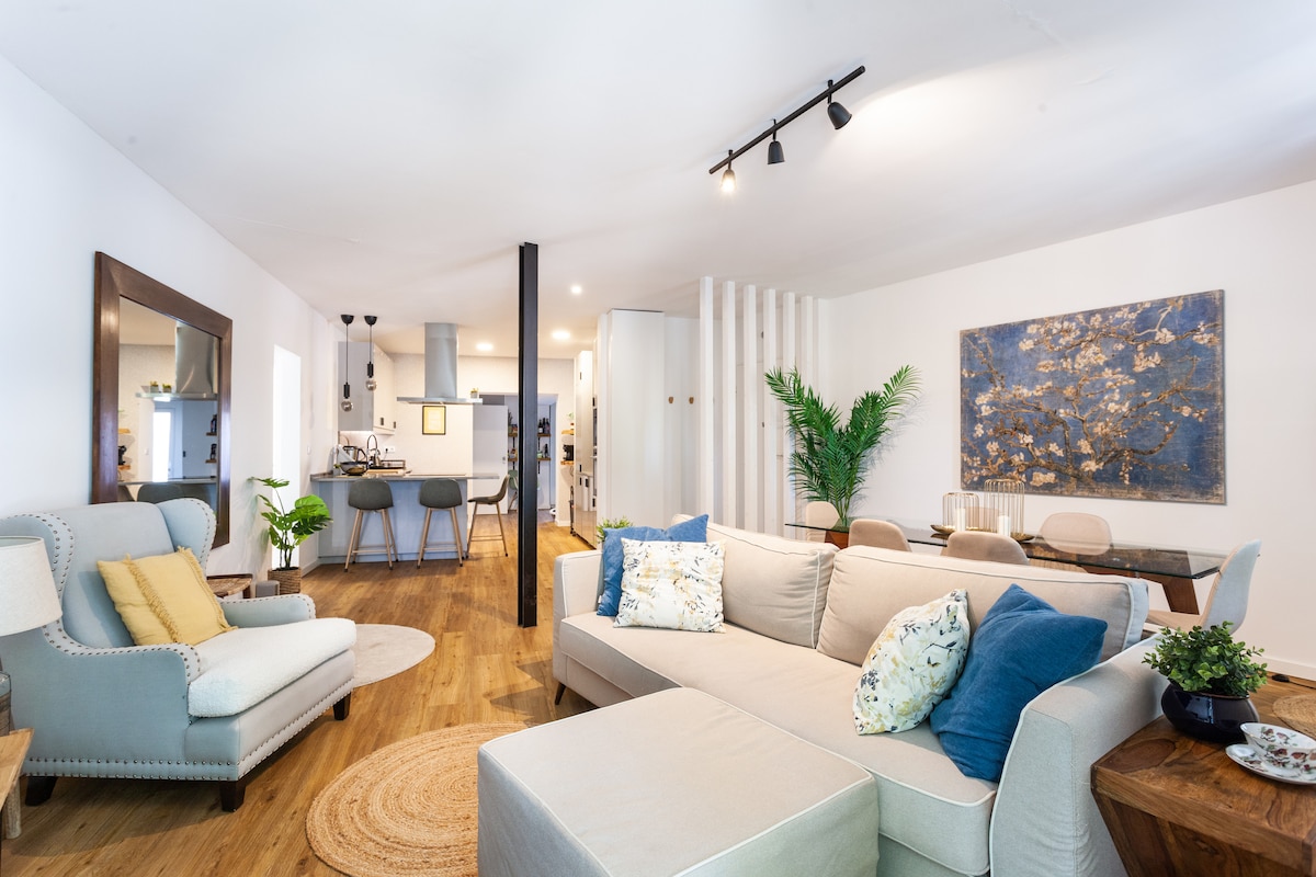 A spacious living area features a large, light-colored sectional sofa adorned with decorative pillows. A round area rug lies beneath the seating arrangement. An elegant dining table accommodates several chairs, while a modern kitchen setup is visible in the background, enhancing the open layout of the space.