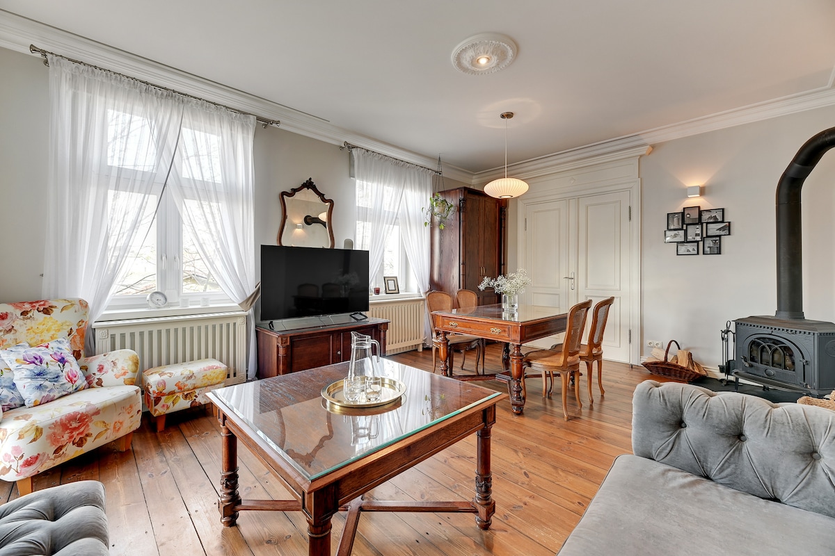 The living area features large windows allowing natural light to fill the space. Comfortable floral-patterned chairs and a plush sofa are arranged around a wooden coffee table. A TV stands on a cabinet, while a dining table is positioned nearby. Decorative elements enhance the inviting atmosphere.