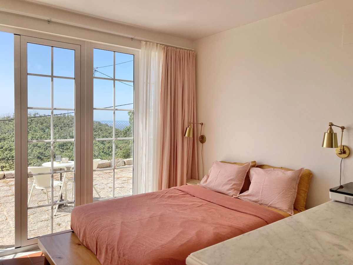 A bright and airy bedroom is seen with large glass doors leading to an outdoor terrace. A spacious bed is made with soft pink bedding, complemented by two bedside wall lamps. Light, airy curtains frame the windows, allowing natural light to fill the room.
