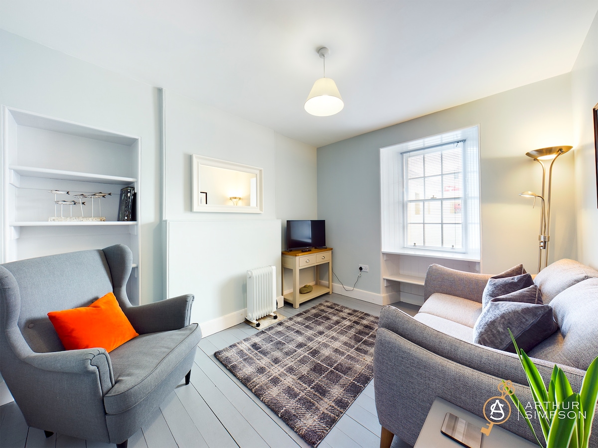A cozy living area is presented with a light grey sofa and a matching armchair, arranged on a textured rug. A television is mounted on the wall across from a window, providing natural light. Shelves display various items, enhancing the functional space.