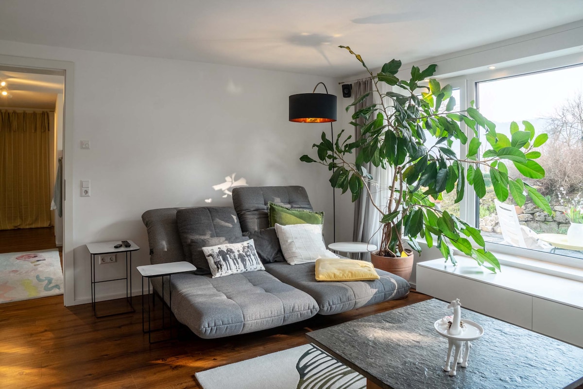 A modern living area is visible, featuring a gray reclining sofa adorned with decorative pillows. A large window allows natural light to fill the space, complemented by a potted indoor plant. A dark coffee table with a decorative piece is positioned in front of the sofa.