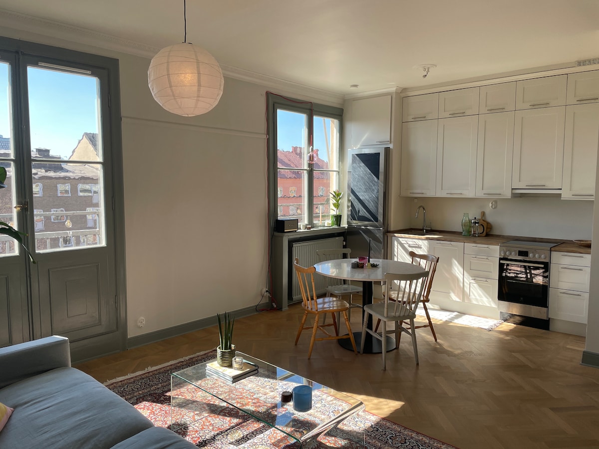 A bright living area features large windows allowing ample natural light. A modern kitchen is visible, showcasing white cabinets and stainless steel appliances. A circular dining table with wooden chairs sits adjacent to a comfortable sofa, enhancing the open and inviting layout.