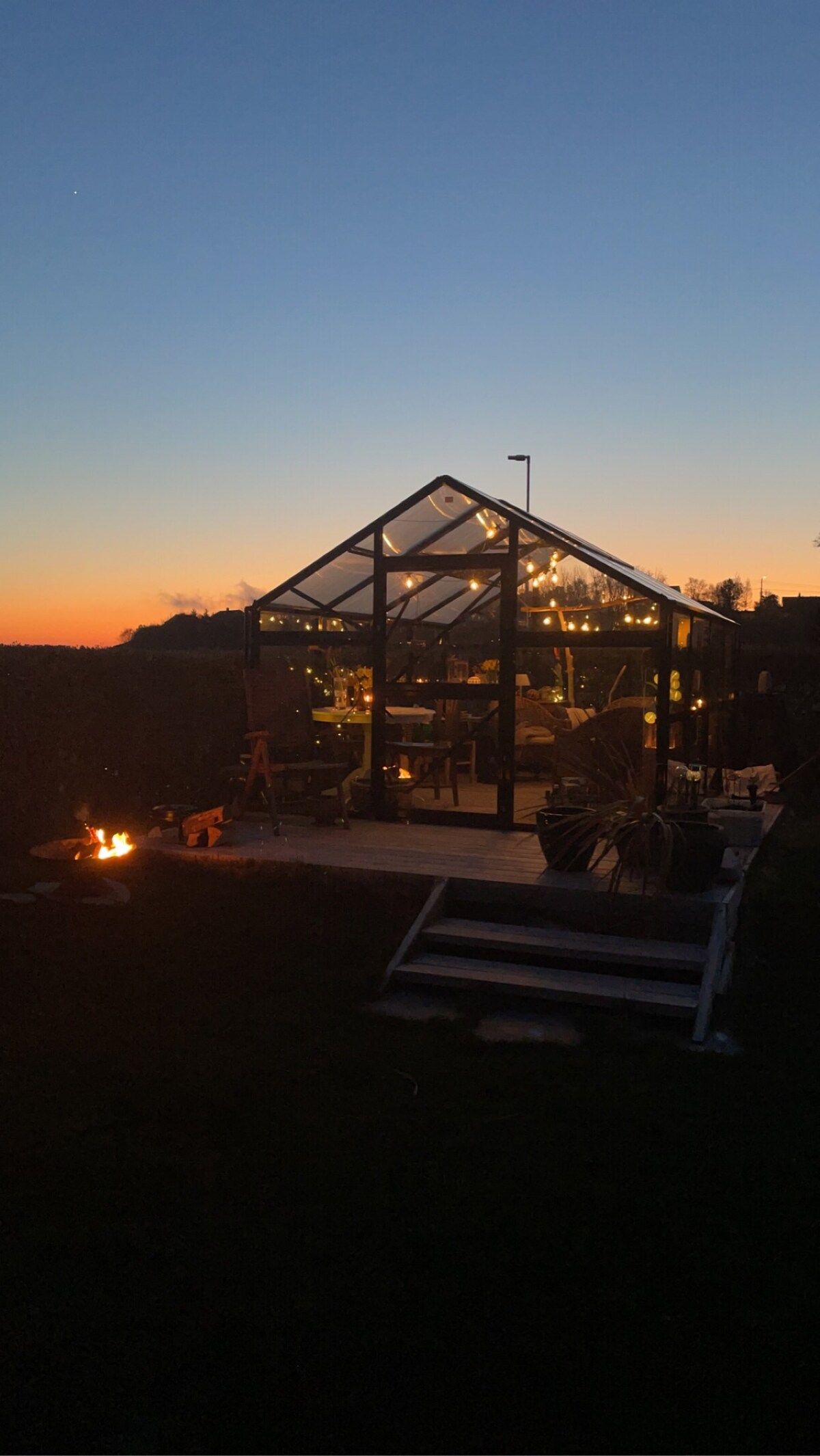A modern greenhouse structure is set against a twilight sky, featuring illuminated interiors visible through the glass walls. A wooden deck surrounds the building, with a firepit glowing nearby, creating a cozy and inviting outdoor space.