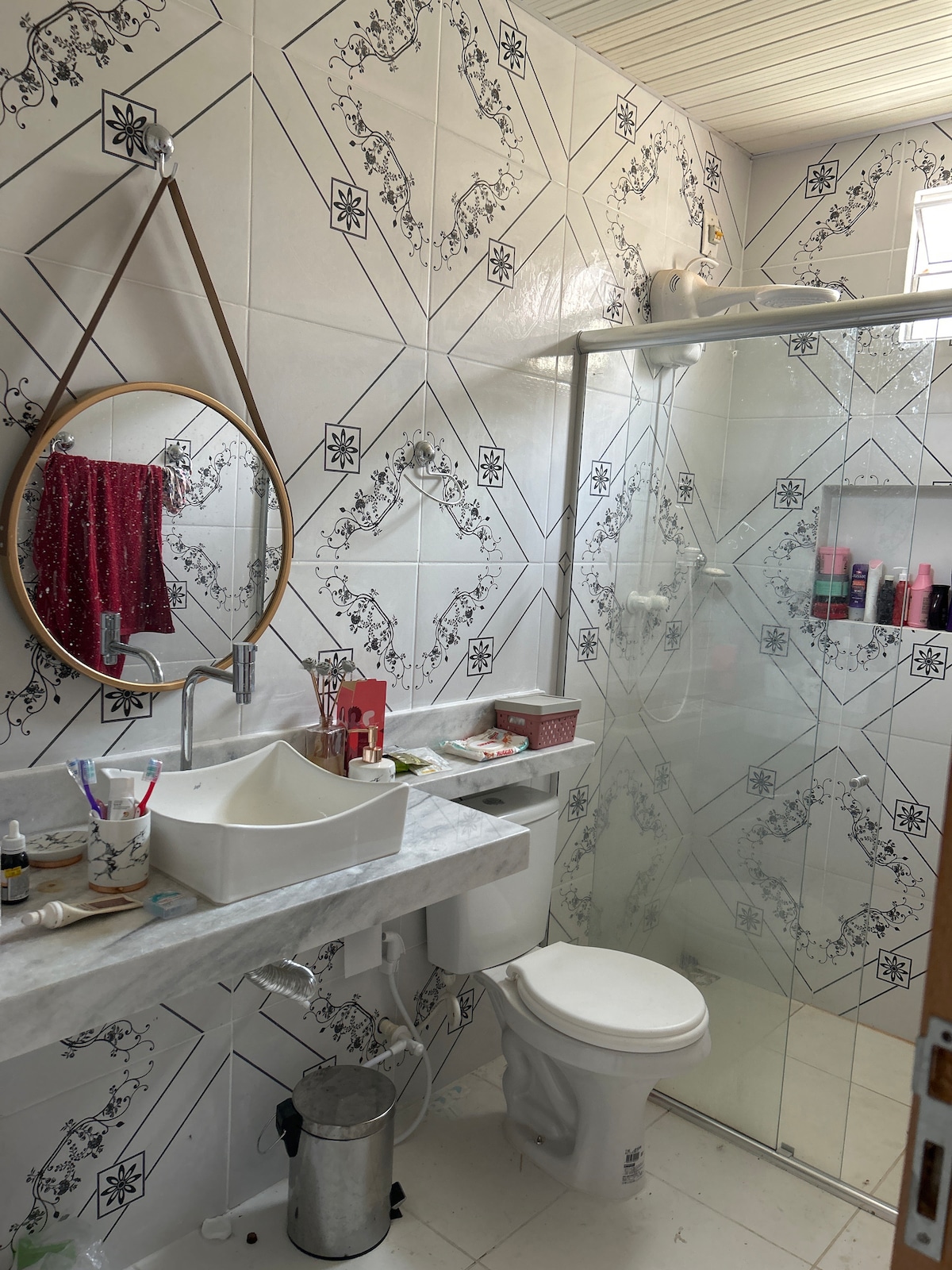 A well-lit bathroom features stylish patterned tiles on the walls. A round mirror hangs above a marble countertop, which is complemented by a white sink. A glass shower enclosure is visible, with toiletries neatly arranged nearby. Natural light enters through a small window.