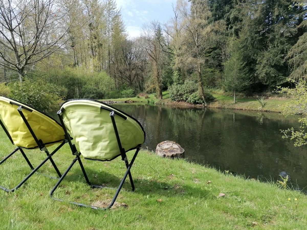 Two green folding chairs are positioned on a grassy bank, overlooking a serene pond surrounded by trees and lush greenery. The tranquil water reflects the surrounding nature, providing a calm setting for relaxation.