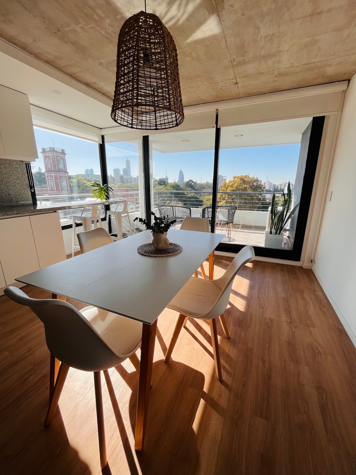 A modern dining area is illuminated by natural light from large windows. A white table is surrounded by four light-colored chairs, and a decorative centerpiece rests on the table. The view showcases city skyline and treetops, enhancing the open atmosphere.