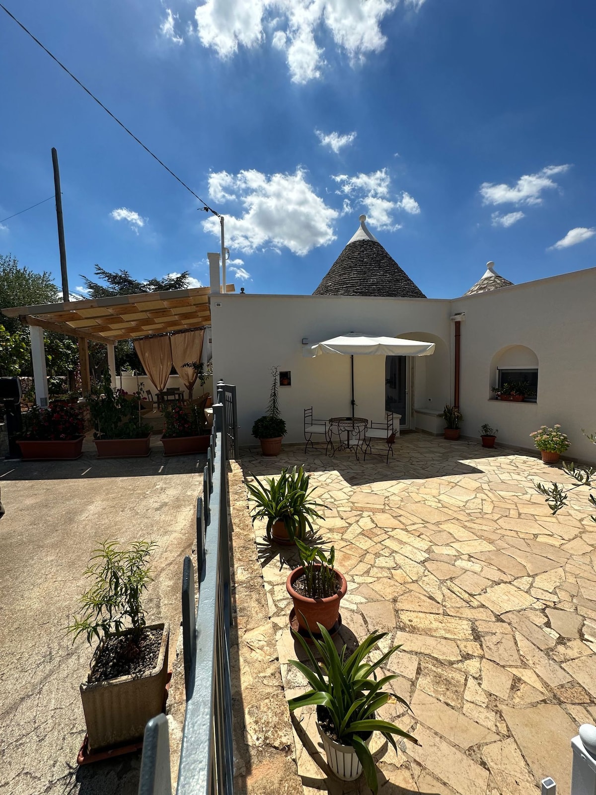 A sunlit outdoor patio features a stone pathway surrounded by potted plants and decorative flowers. A small table with two chairs sits beneath a shaded umbrella. The structure showcases traditional trullo architecture with conical roofs, complemented by a pergola draped with fabric.