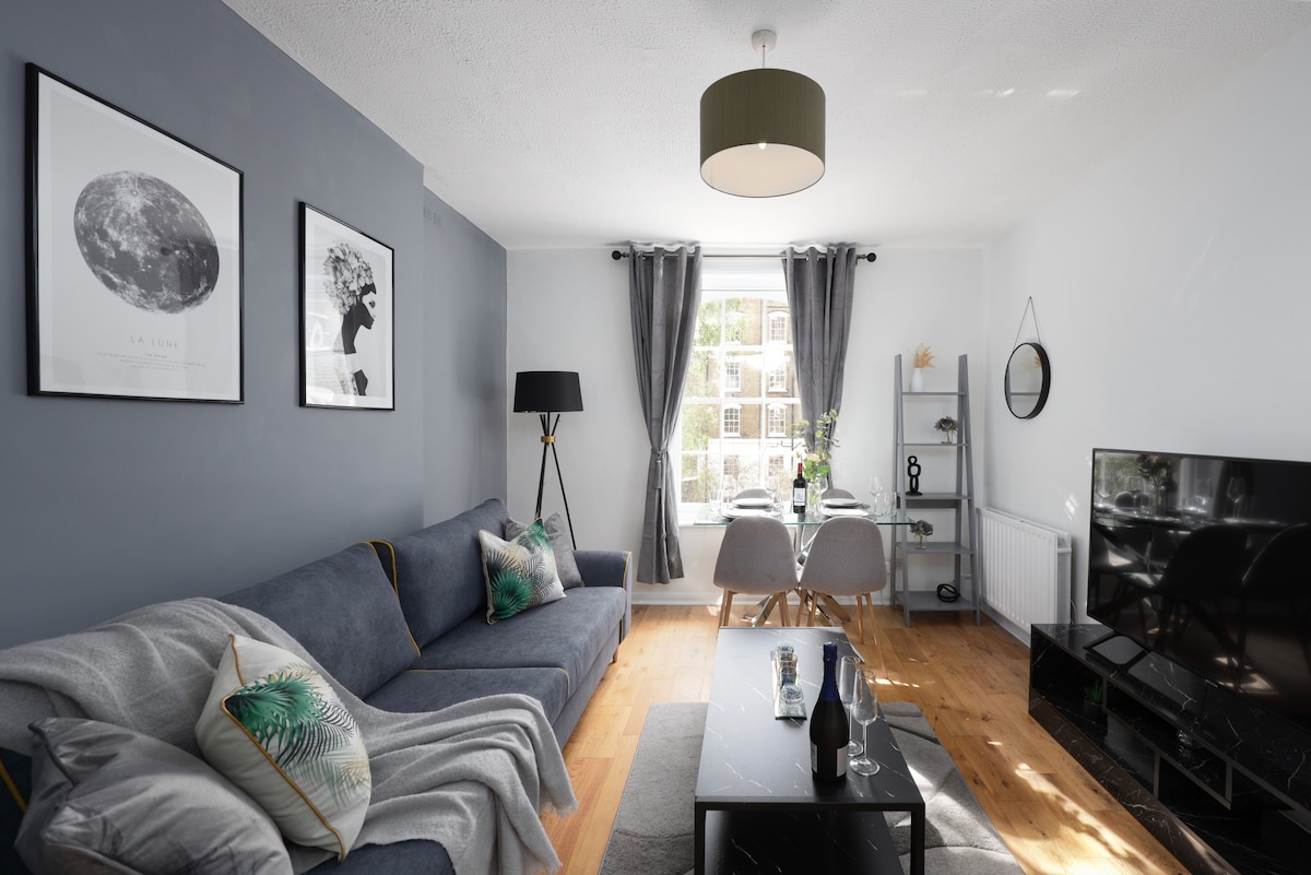 A modern living room offers a bluish-gray sofa adorned with decorative cushions. A round dining table for four is positioned near the window, allowing natural light to brighten the space. Artwork is displayed on the walls, and a large television is situated on a sleek black media console.