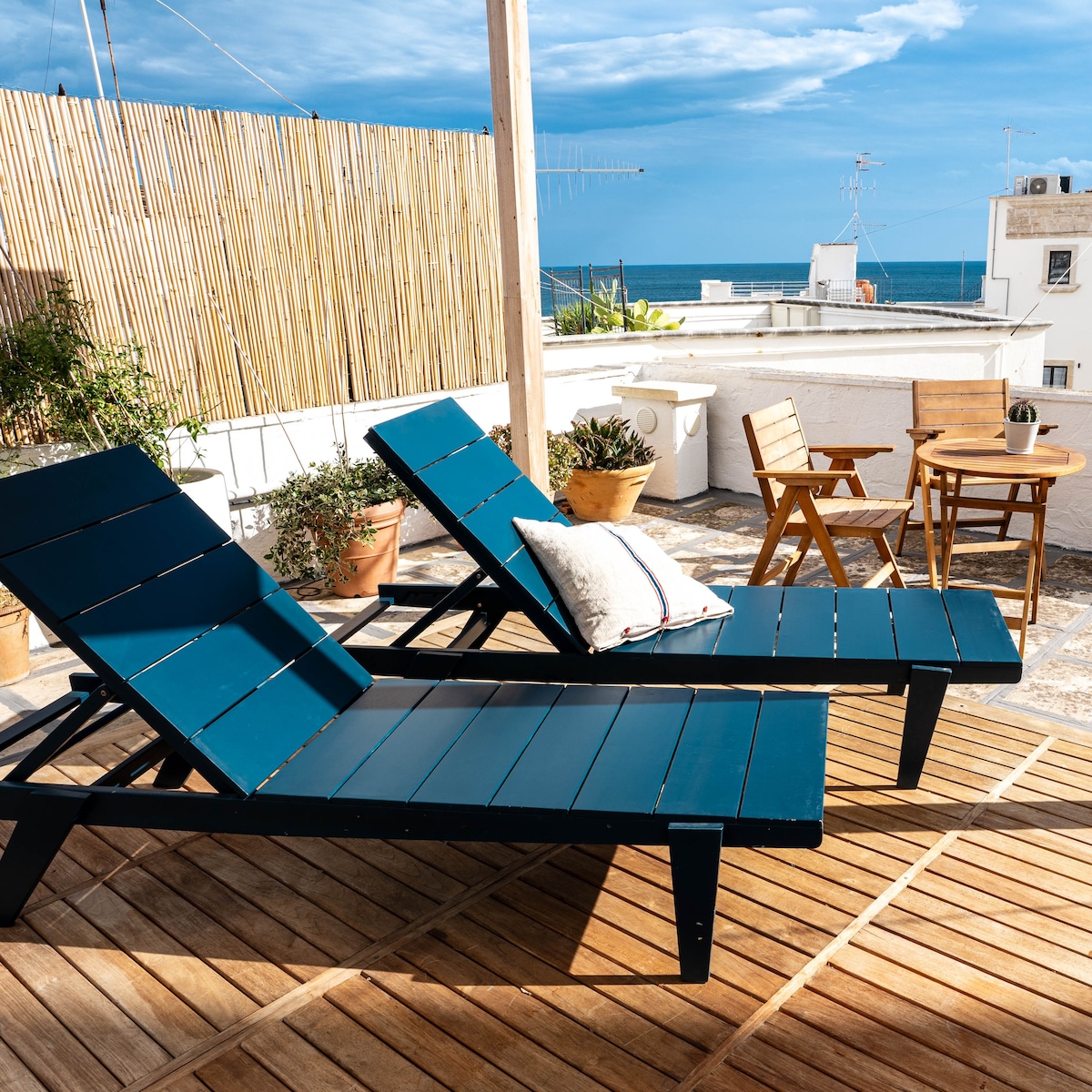 The spacious terrace features two modern lounge chairs in deep blue, positioned for ocean views. A small wooden table with two matching chairs is nearby. Potted plants add greenery, while a bamboo fence provides privacy against the sky and sea backdrop.