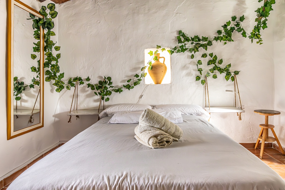 A cozy sleeping area features a spacious bed adorned with neatly stacked towels. Shelves with decorative elements are arranged beside the bed, while a mirror reflects the bright, neutral walls. Greenery drapes elegantly along the wall, complemented by a window showcasing an earthenware vase.