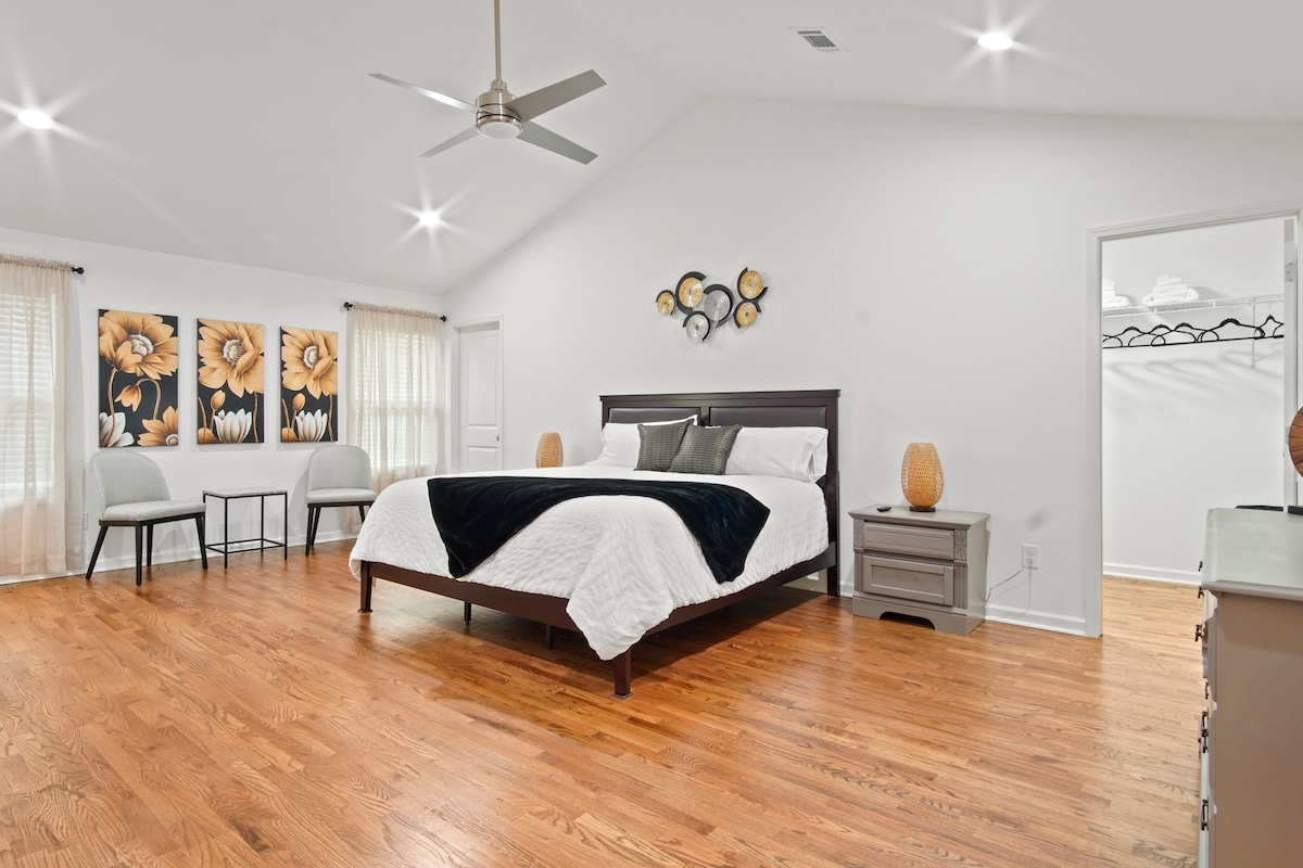 A spacious master bedroom features a large bed with crisp linens, accompanied by two chairs and a small table. Soft lighting illuminates the room, highlighting the elegant flower artwork on the wall. A walk-in closet is accessible in the background.