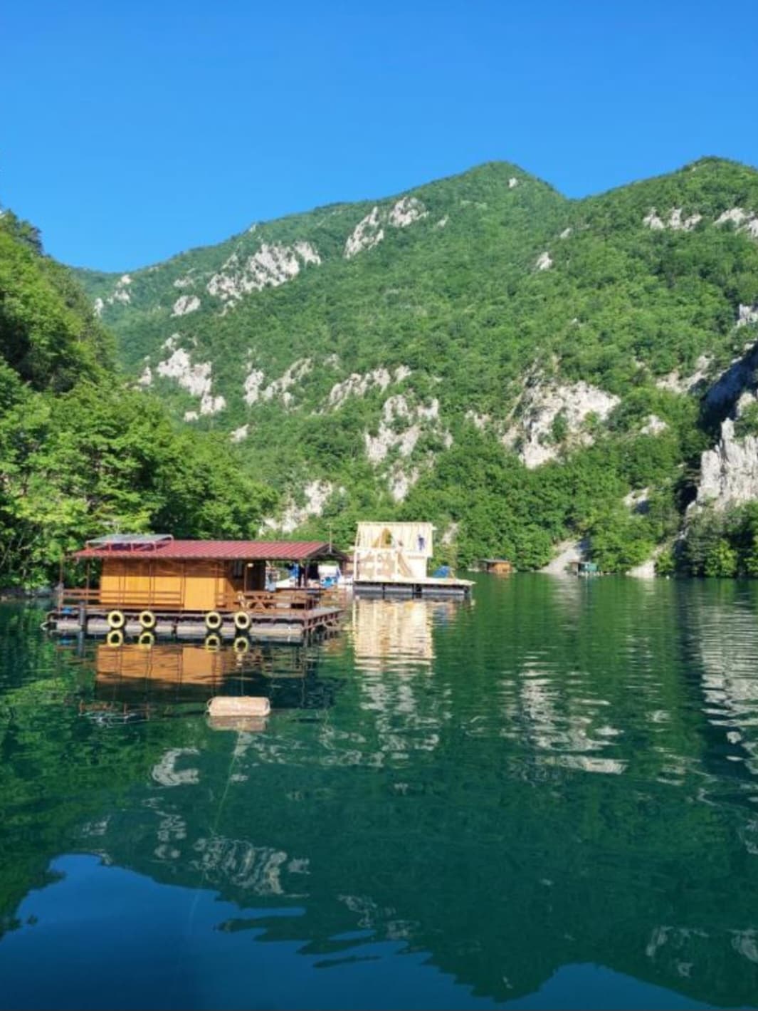 A serene view of the floating apartments on clear waters, surrounded by lush green mountains. The reflections of the vibrant landscape can be seen in the calm surface of the water, complemented by a sunny, blue sky above.