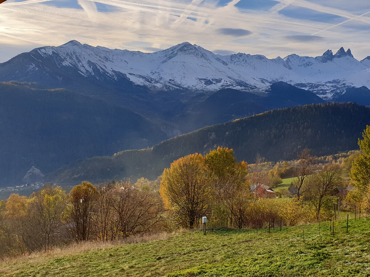 The image shows a panoramic view of snow-capped mountains under a bright sky with crossing clouds. Lush valleys and trees with autumn foliage are visible in the foreground, showcasing a vibrant contrast with the icy peaks in the distance.