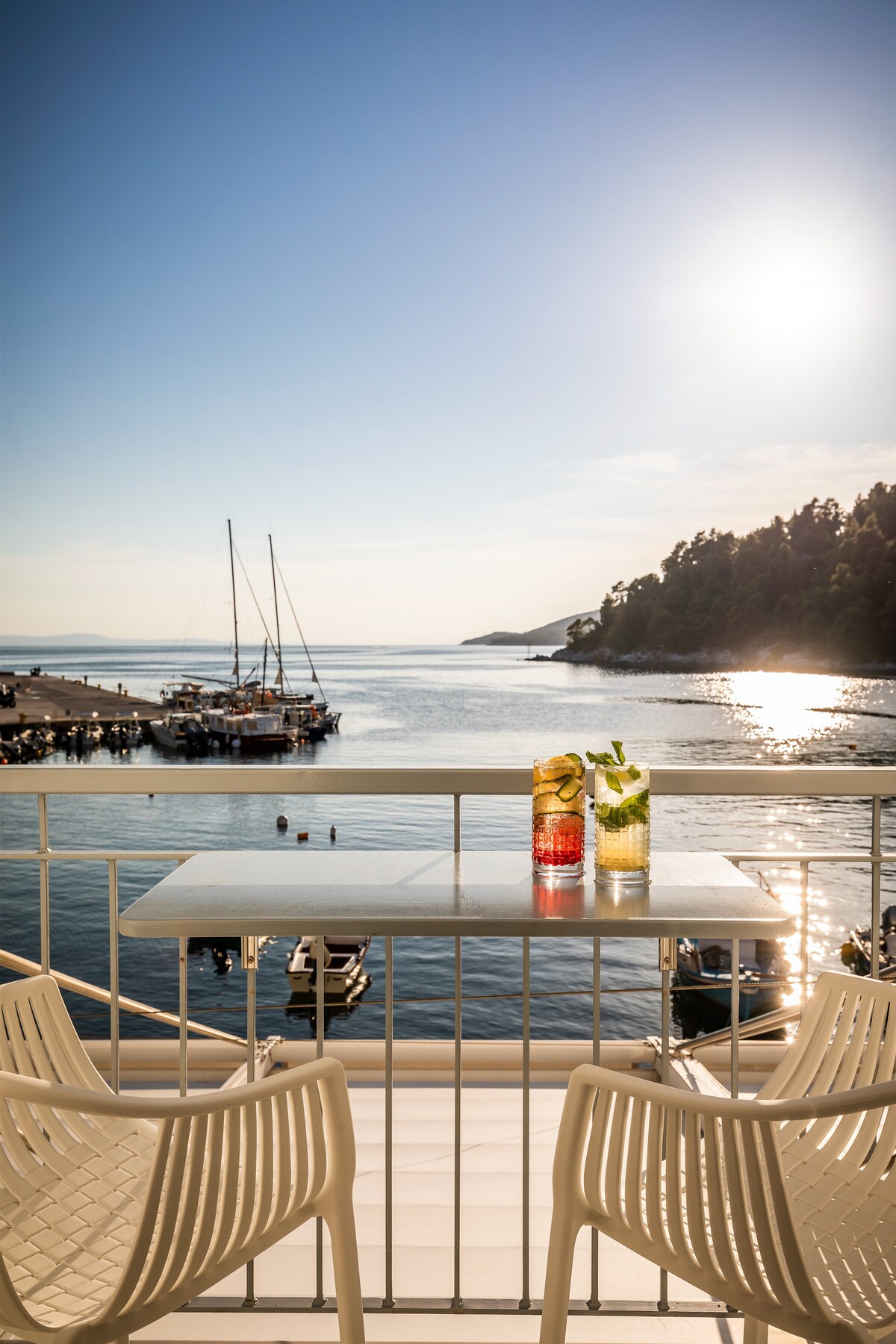 A private balcony features a table with two drinks, overlooking the serene bay and boats in the water. The sun sets in the background, casting a warm glow over the scene. Two chairs are positioned at the table, inviting relaxation with a scenic view.