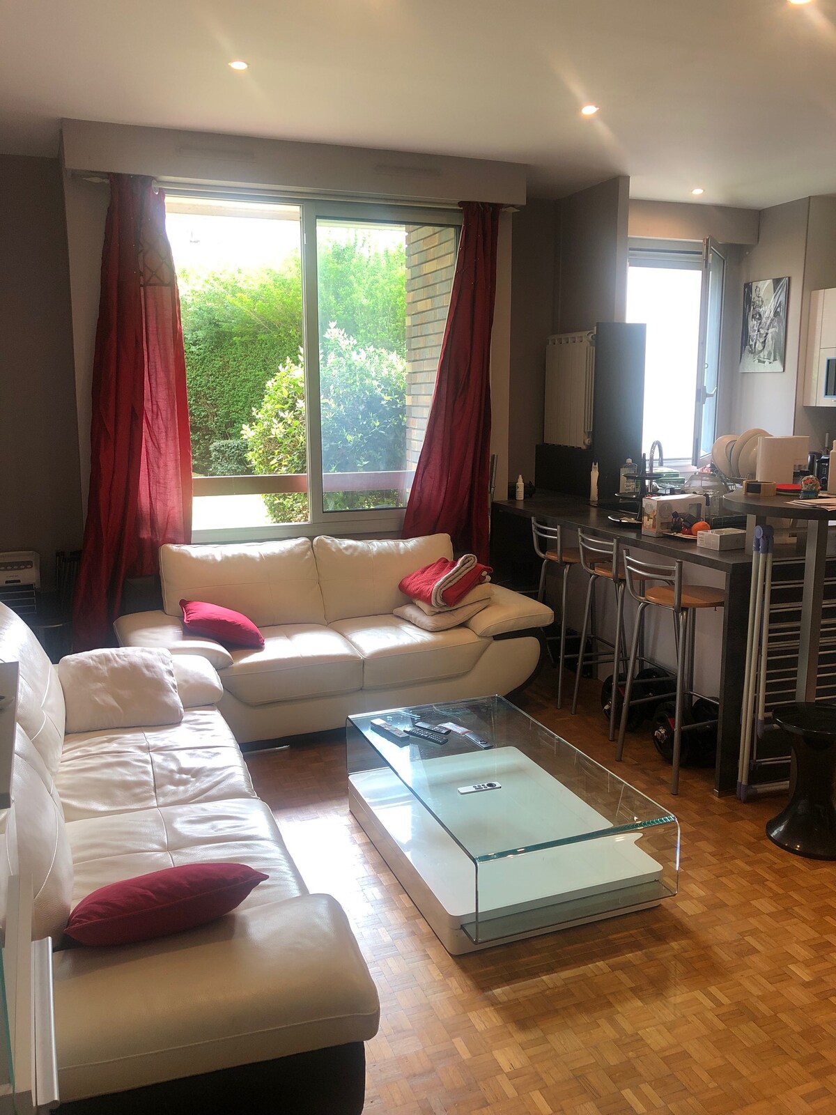 A bright living area features a white leather sofa adorned with colorful cushions and a clear coffee table. Large windows bring in natural light and showcase greenery outside. A bar area with high stools is positioned next to the kitchen space.