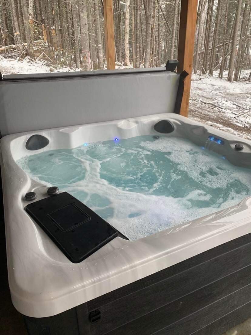A modern hot tub is situated under a covered structure, surrounded by trees. Bubbles rise gently from the water, accented by soft blue lighting. The tub is designed for relaxation, providing a tranquil escape in a natural setting.