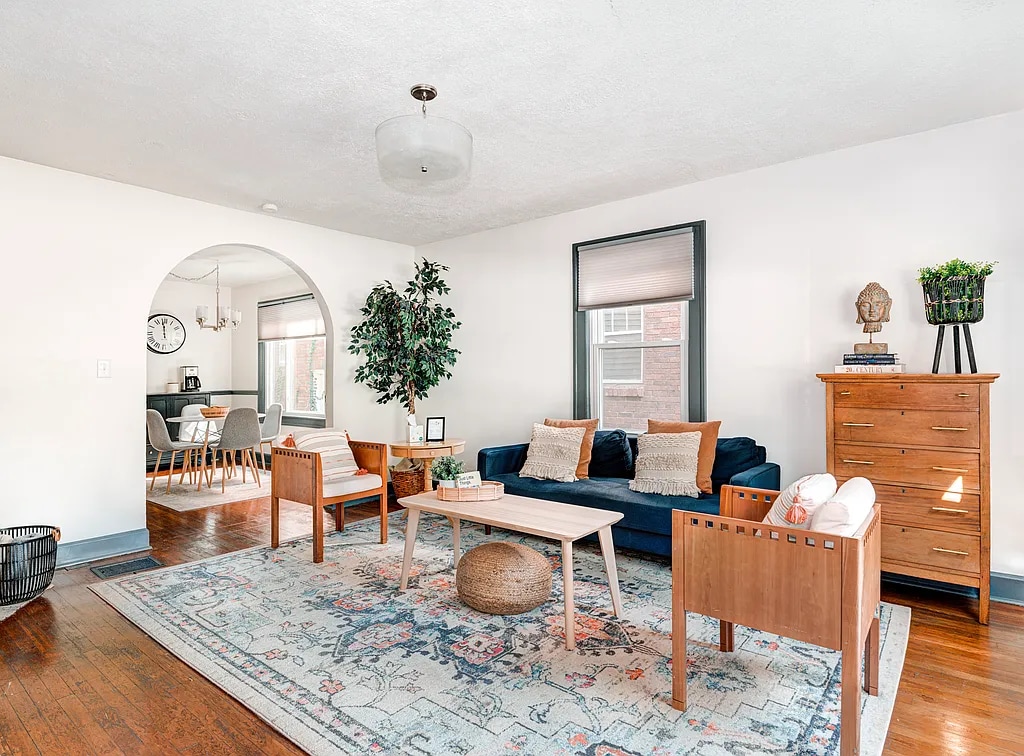 A spacious living area features a deep blue sofa centered on a light area rug. Two wooden armchairs are arranged beside a wooden coffee table. A tall plant adds greenery, and a dresser with decorative items stands against the wall, illuminated by natural light from a large window.