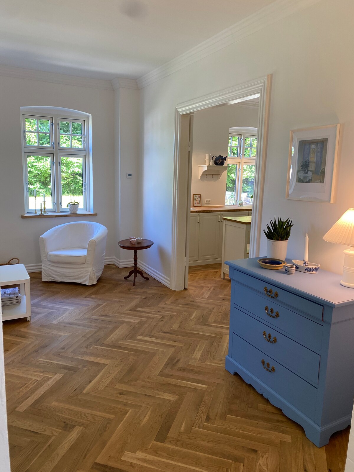 A bright entrance area features a light-colored chair beside a small side table. The warm wood flooring complements a blue dresser. Natural light streams through the multi-paned window, enhancing the inviting ambiance. A doorway leads to the kitchen area visible in the background.
