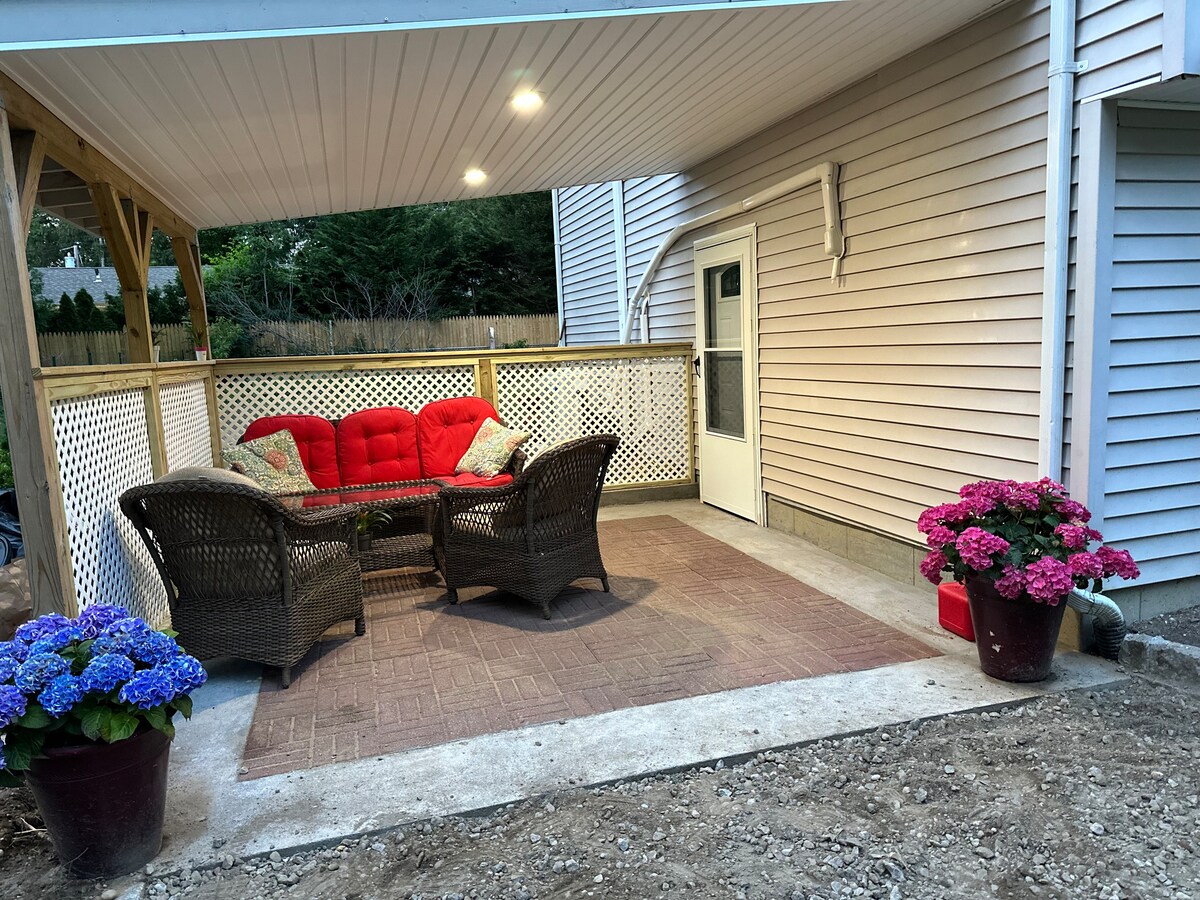 A covered outdoor patio offers a cozy seating area with a red sofa and two wicker chairs. Brightly colored flower pots filled with blue and pink blooms are placed on either side, enhancing the inviting atmosphere of the space.