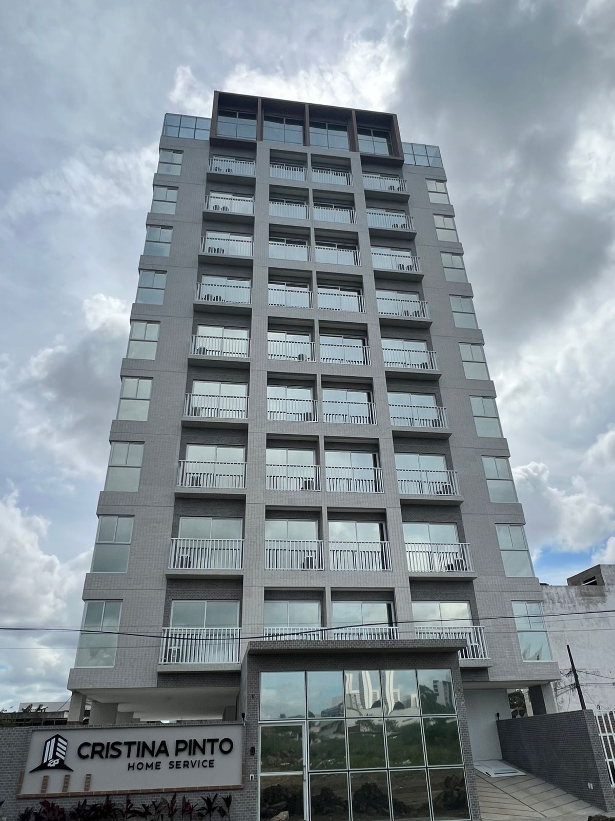 A modern apartment building features a sleek facade with multiple balconies and large windows. The entrance is marked by a glass front, providing an inviting view of the lobby area. The sky above is partly cloudy, adding to the building's contemporary appeal.