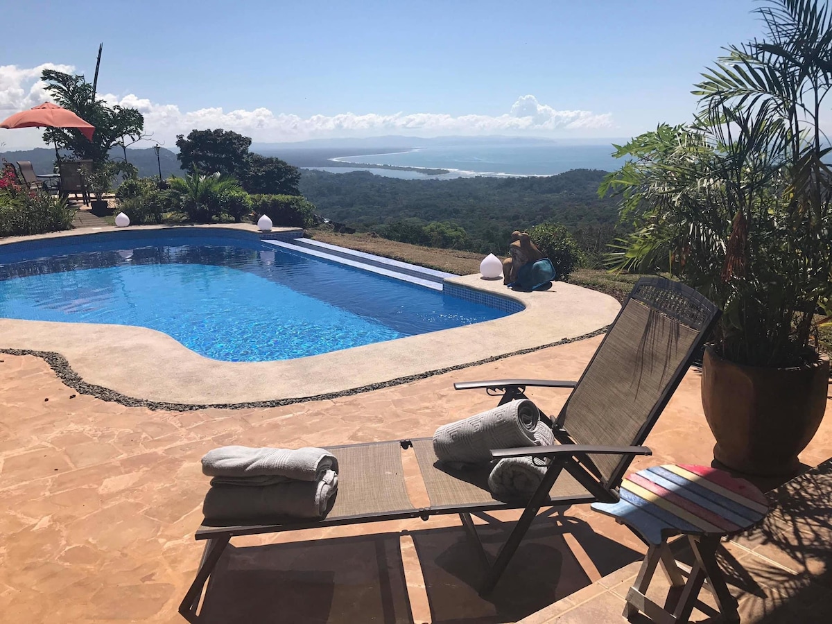 A serene outdoor pool area is visible, framed by lush greenery and offering expansive ocean views. A reclining lounge chair is positioned alongside the pool, with neatly rolled towels placed on it. Bright sunlight reflects off the water, enhancing the relaxing atmosphere.