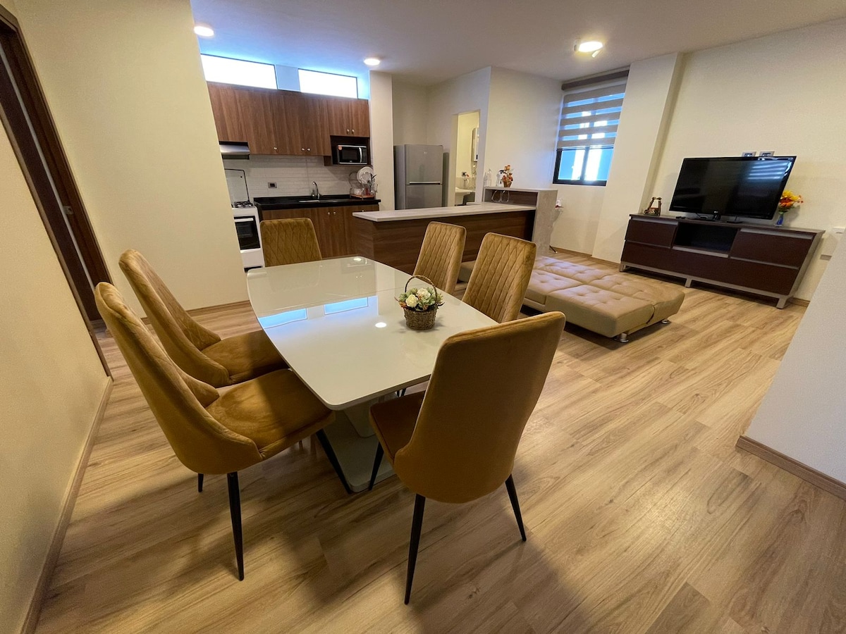 A modern dining area includes a sleek table surrounded by four upholstered chairs. A beige sofa bed is positioned adjacent to the table, with light wood flooring extending throughout. The kitchen area features contemporary cabinetry and stainless-steel appliances, offering an open layout.