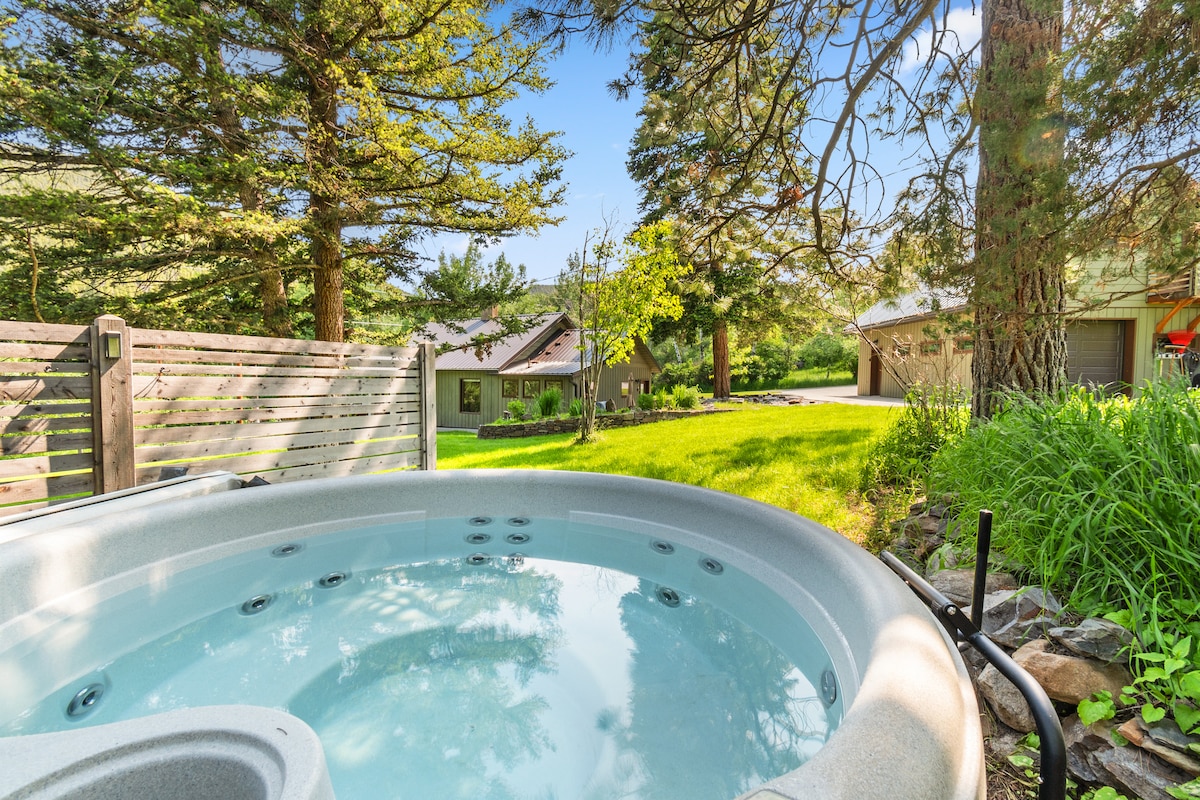 A hot tub is positioned in a lush yard surrounded by tall trees, with a view of a nearby house and the driveway. The clear water reflects the blue sky, inviting relaxation in a serene outdoor setting.