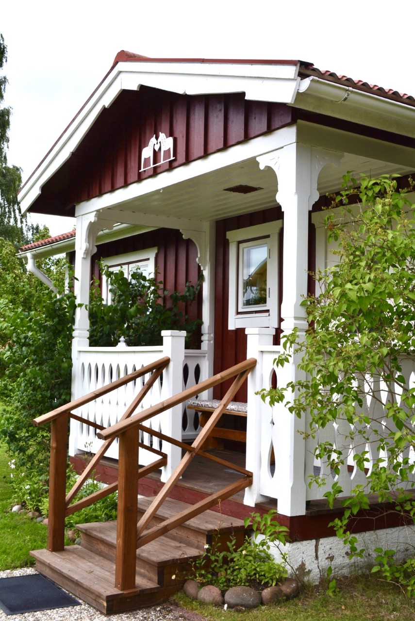 A charming entrance features a wooden staircase leading up to a door framed by decorative white trim. The façade is painted in a rich red, complemented by lush greenery on either side, creating an inviting approach to the house.