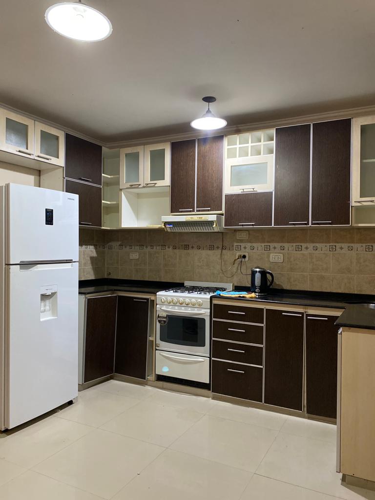 A modern kitchen features dark wooden cabinetry contrasted by light countertops. Essential appliances, including a refrigerator and a stove, are present. Ample storage is provided through upper cabinets with glass panels. A neutral tile floor complements the overall design.