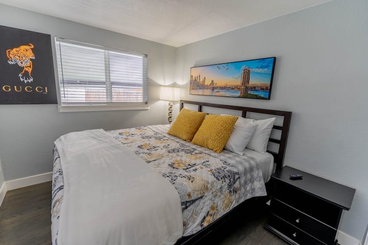 Тази спалня разполага с голямо двойно легло със стилна табла, акцентирано с шарено спално бельо и ярки жълти възглавници. Нощното шкафче и подовата лампа осигуряват функционалност, а произведенията на изкуството и неутралните стени подчертават модерния, приветлив дизайн.