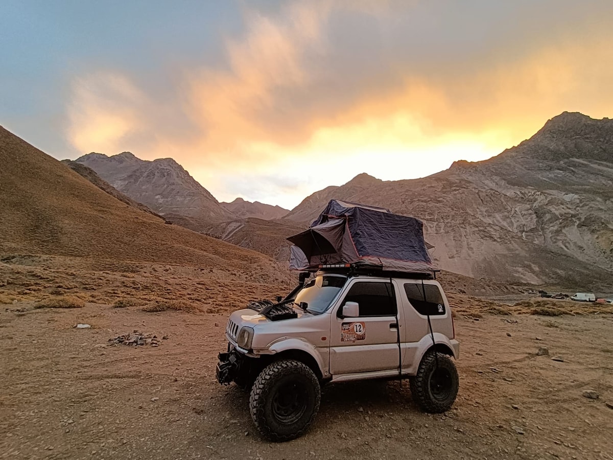 A compact gray 4x4 vehicle is parked on a rugged terrain under a dramatic colorful sky at sunset. A rooftop tent is visible, providing additional sleeping space. Surrounding mountains showcase earthy tones, highlighting the natural beauty of the landscape.