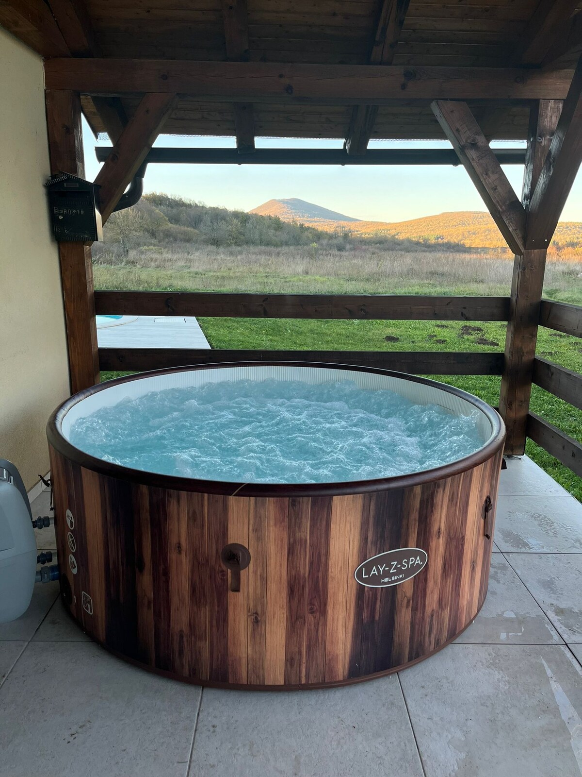 A wooden hot tub is situated on a patio area, surrounded by a railing and offering views of rolling hills. The bubbling water is visible, while a mountain peak rises in the background under a clear sky.