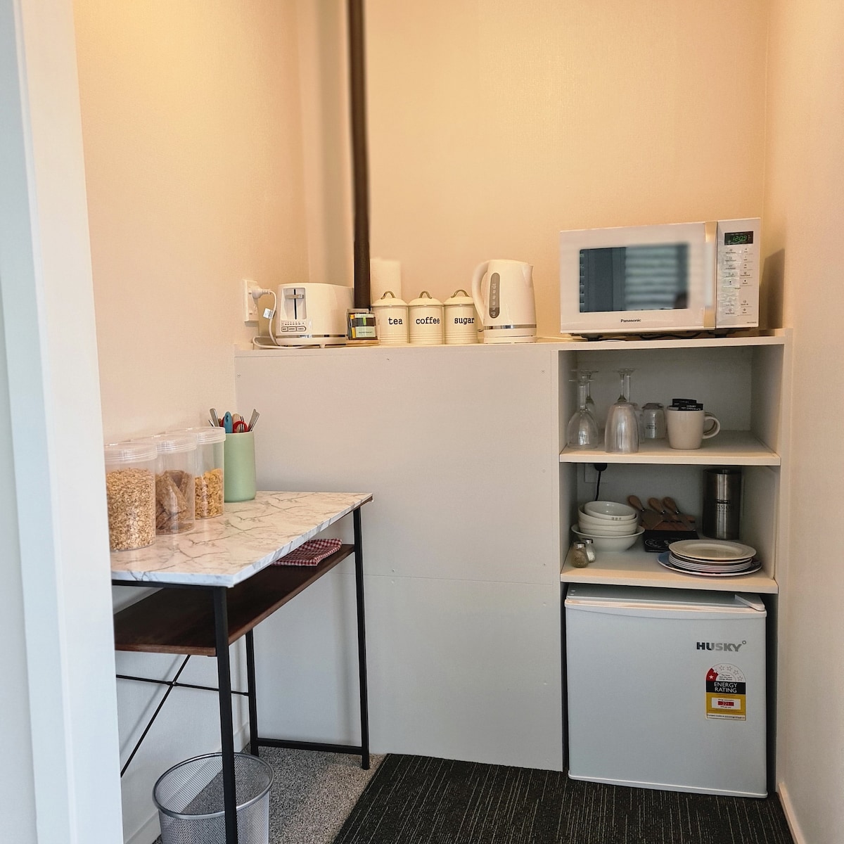 A compact kitchenette is equipped with a microwave, kettle, and storage containers labeled for tea and coffee. A small table holds jars of cereals and utensils, while a mini-fridge is positioned below. Light-colored walls brighten the space, creating a functional cooking area.