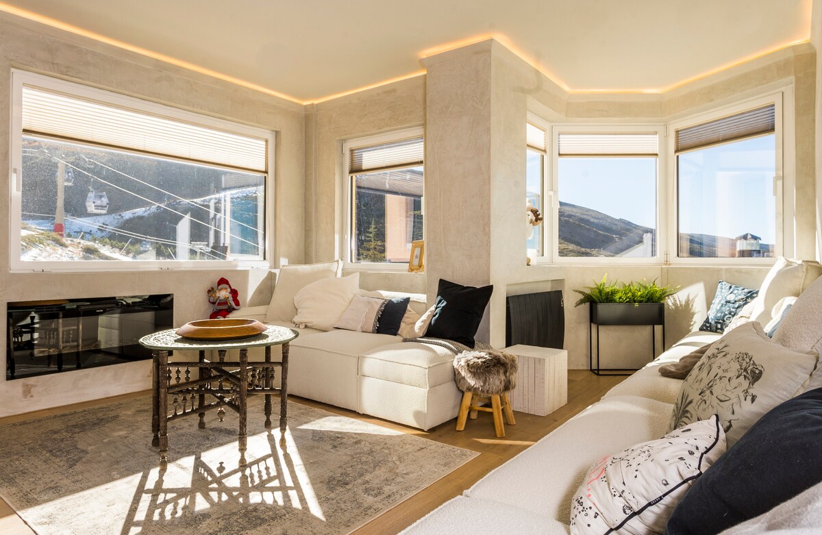 A spacious living area features an L-shaped sofa positioned around a central coffee table. Large windows provide abundant natural light and offer panoramic views of the surrounding slopes. A gas fireplace contributes to the cozy atmosphere, complemented by decorative pillows and a plant.