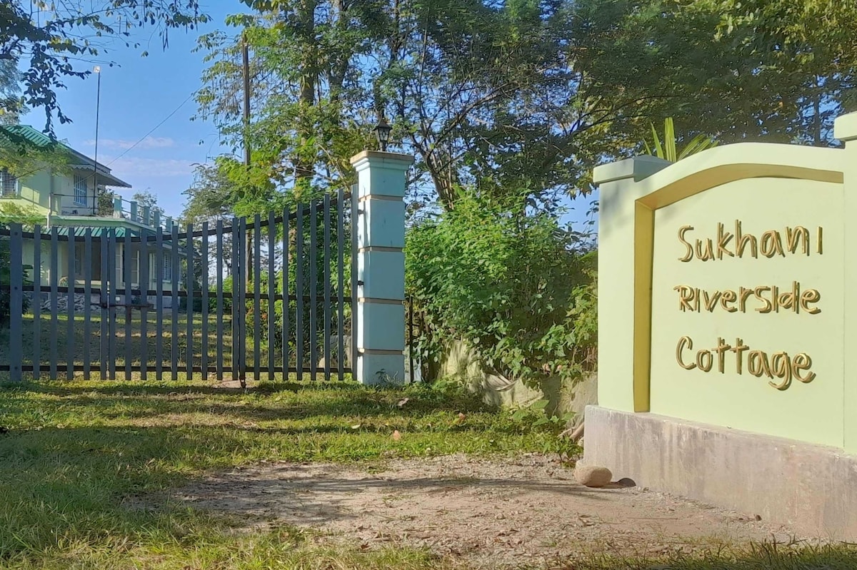 The entrance to Sukhani Riverside Cottage is depicted, featuring a welcoming sign set against a lush green landscape. A wooden gate provides access to the property, framed by trees and open grass, leading towards the charming cottage situated in the background.