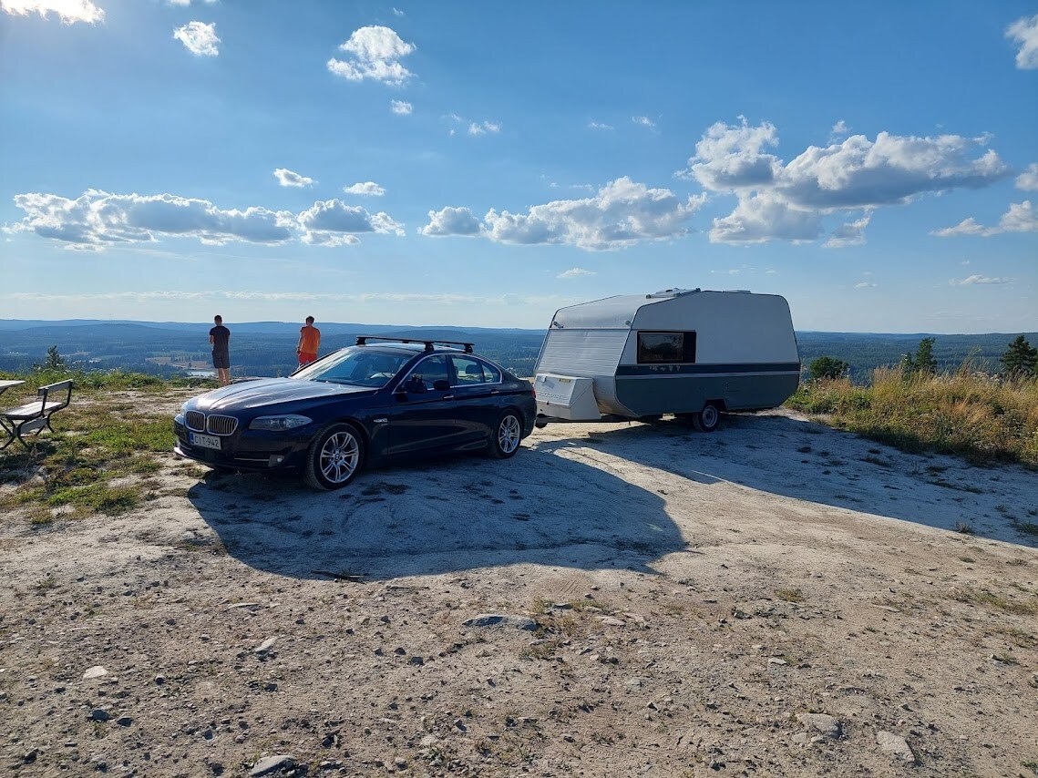 A car is parked next to a caravan set against a scenic view. The landscape features rolling hills under a blue sky with scattered clouds. A picnic table is visible nearby, offering a place to relax and enjoy the surroundings.