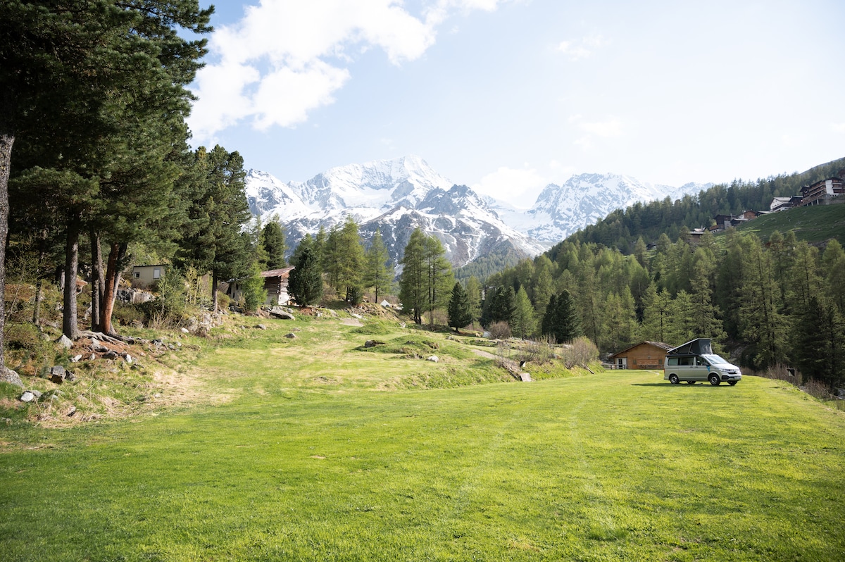 A spacious green area is surrounded by tall trees, with stunning mountainous landscapes in the background. The sky is clear, showcasing a mix of blue and white clouds. A vehicle is parked on the grass, suggesting accessibility and convenience.