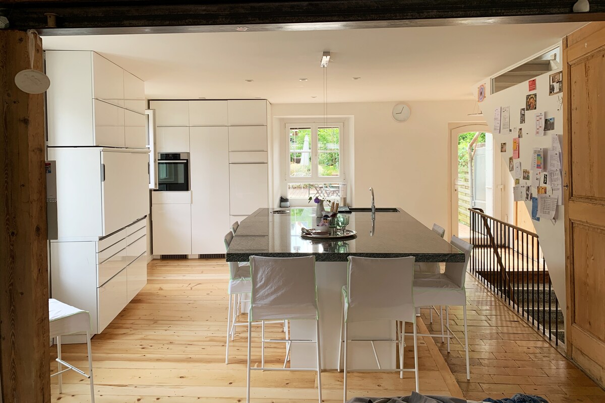 A modern kitchen showcases sleek, white cabinetry and an expansive central island with seating. Natural light fills the space through a nearby window, highlighting the warm wooden flooring. A spiral staircase is visible, leading to additional living areas.