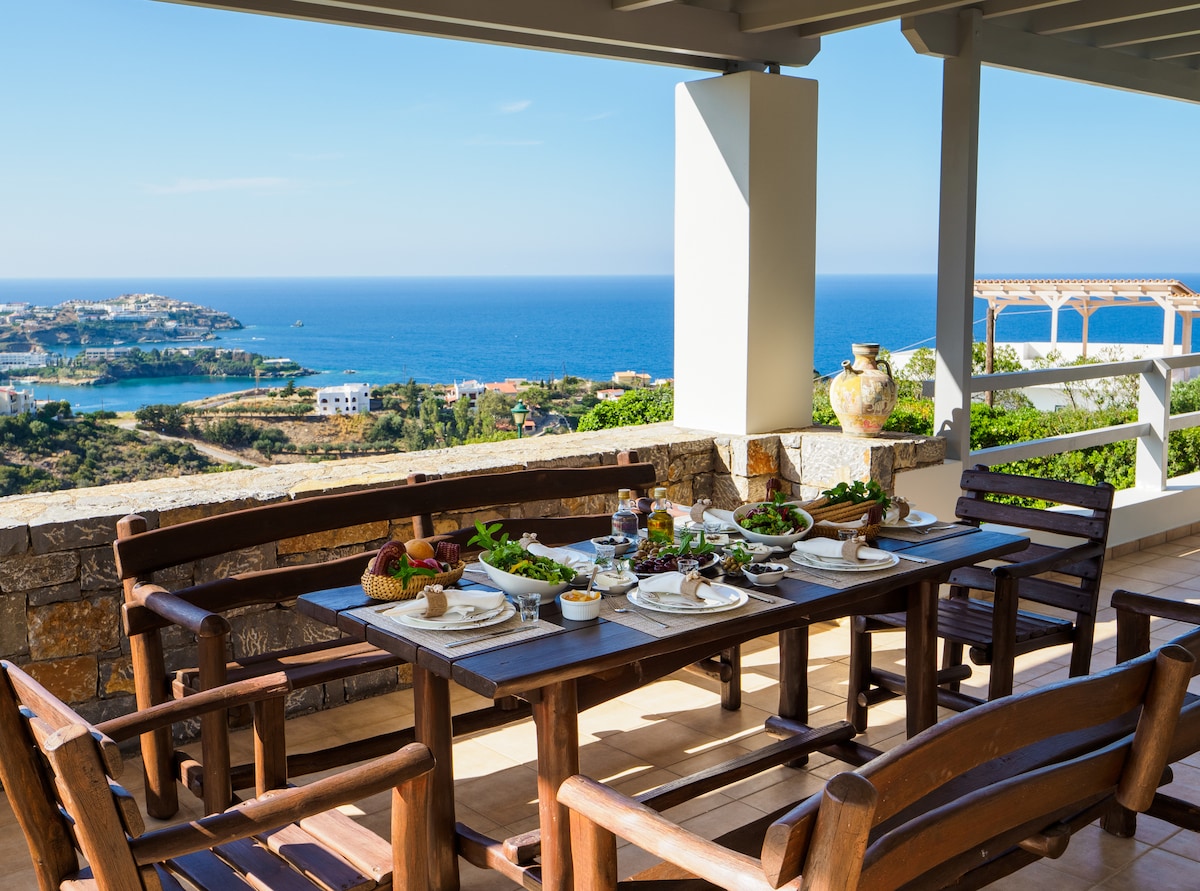 An outdoor dining area is set on a spacious terrace, featuring a wooden table surrounded by sturdy chairs. The table is arranged with plates of food and fresh ingredients. A panoramic view of the sea and surrounding landscape is visible in the background.