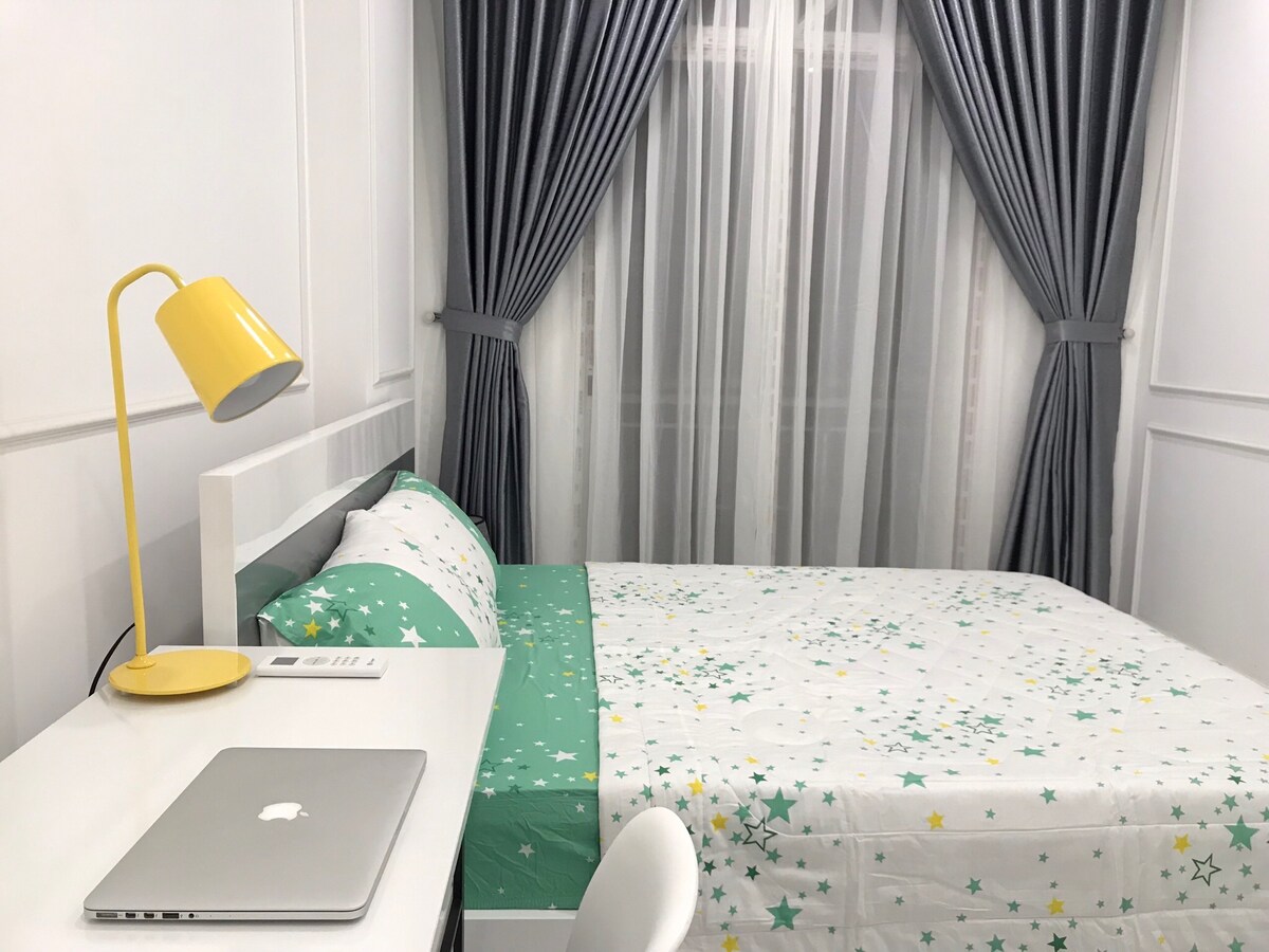A well-lit sleeping area features a spacious bed with a vibrant star-patterned comforter. A sleek desk with a yellow lamp is positioned beside the bed, while gray curtains frame a window allowing natural light to enter.
