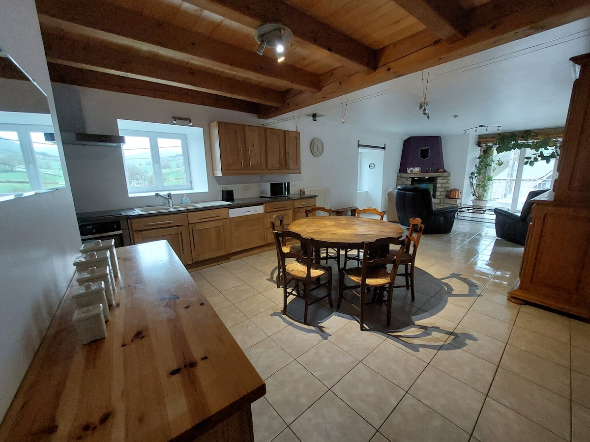 The kitchen area features a rustic design with wooden cabinetry and a round dining table surrounded by wooden chairs. A spacious countertop offers ample space for meal prep, while a large window allows natural light to fill the room, revealing views of the surrounding landscape.