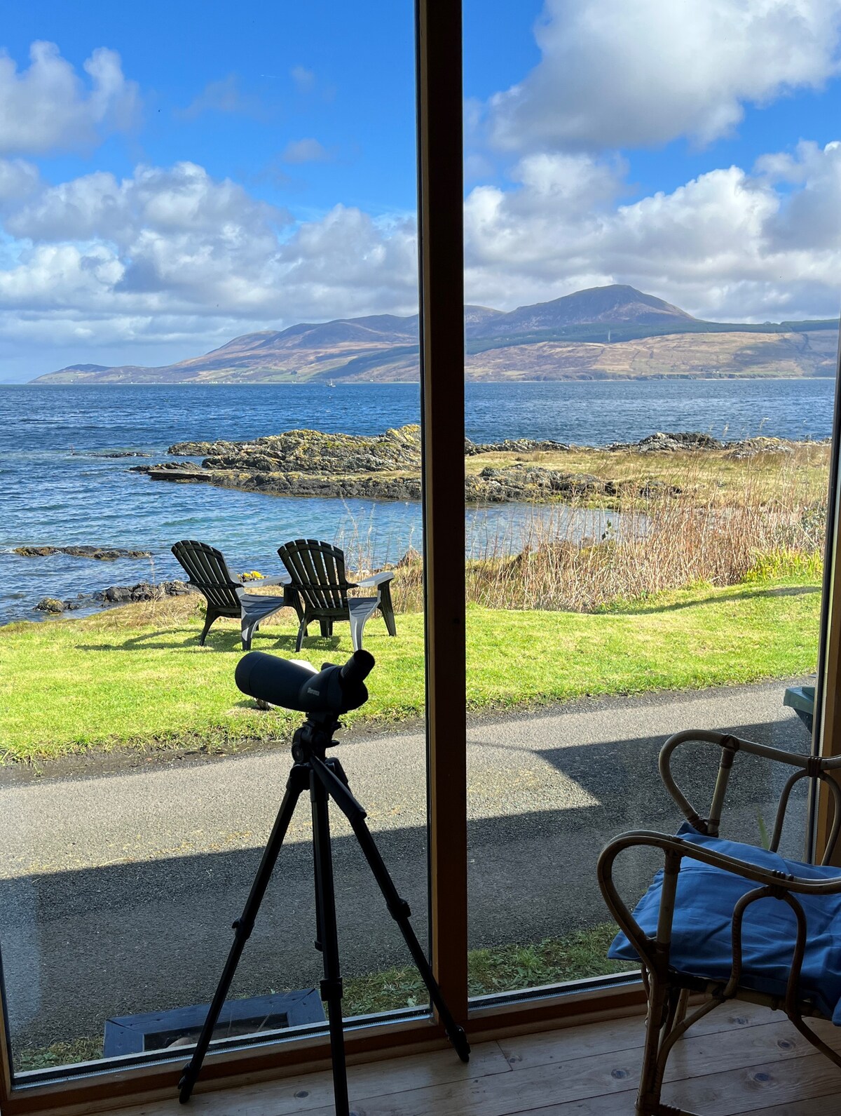 A large window frames a scenic view of the coastline, showcasing rolling hills and a vibrant sky. Two outdoor chairs are positioned on the grass outside, accompanied by a telescope on a tripod, ready for watching wildlife and surveying the landscape.