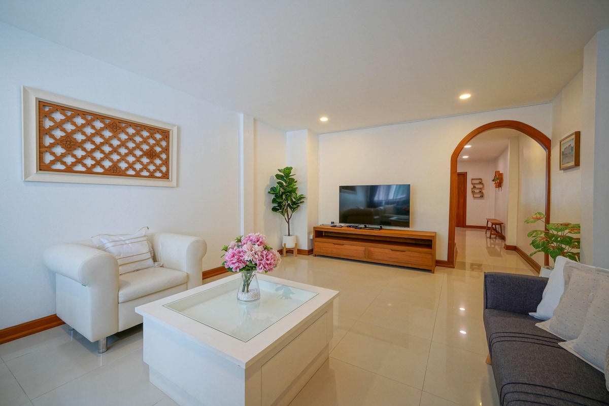 A bright living area is presented, featuring a modern white couch and a dark sofa with decorative cushions. A coffee table with a floral arrangement sits in the center. A media console is positioned against the wall, with a flat-screen television above it, and ambient lighting enhances the space.