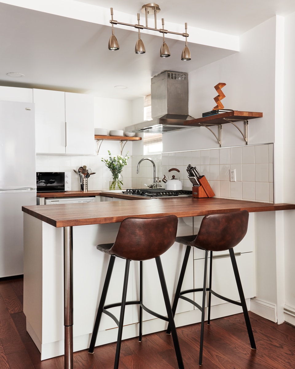 The kitchen area features a modern bar with two high-profile chairs and a polished countertop, complemented by sleek white cabinetry. A stainless-steel vent hood is positioned above the stove. Natural light filters through a nearby window, enhancing the inviting space.
