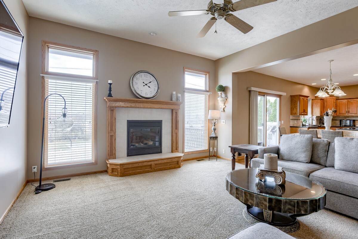 A spacious living room features large windows allowing natural light to fill the area. A cozy gas fireplace is framed by light-colored wood, accompanied by a wall clock and decorative lamps. A circular coffee table is positioned in front of a large sectional couch.
