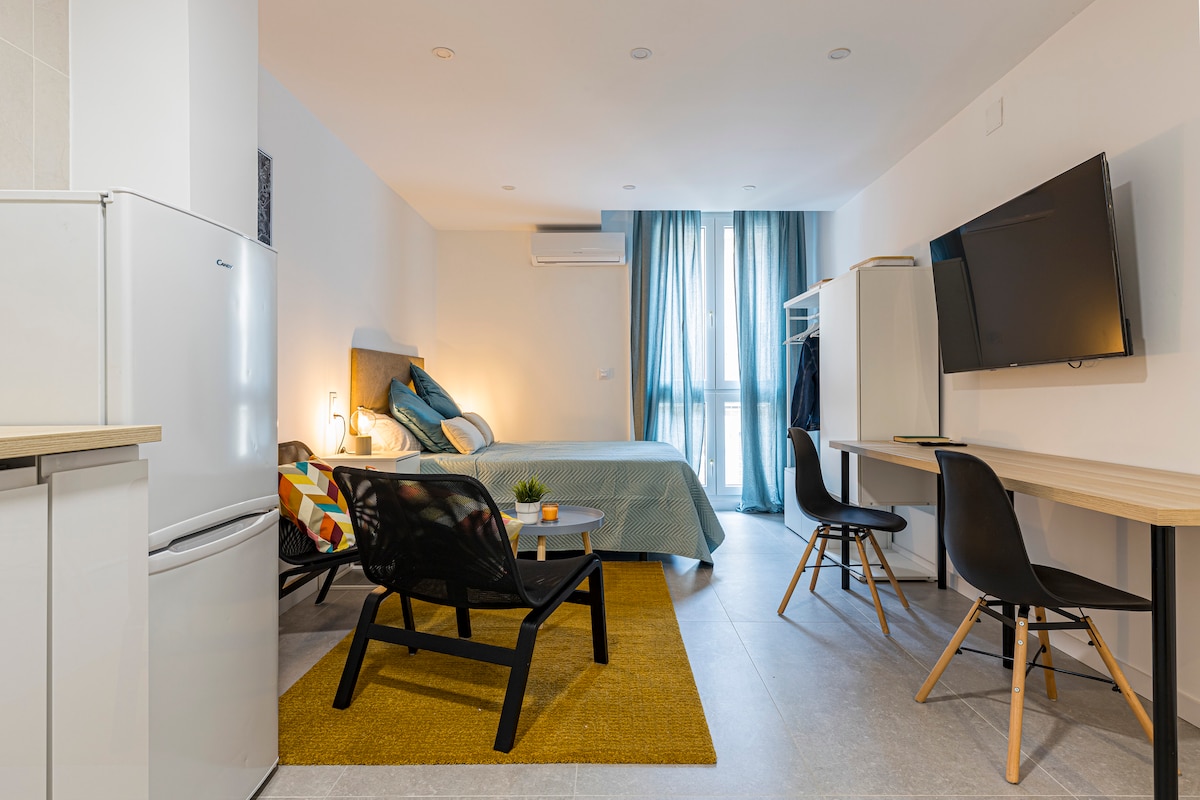 A bright studio apartment is showcased with a spacious bed in a light color scheme. A comfortable seating area is visible, alongside a small dining table and chairs. Large windows are draped with light fabric, allowing natural light to fill the space.
