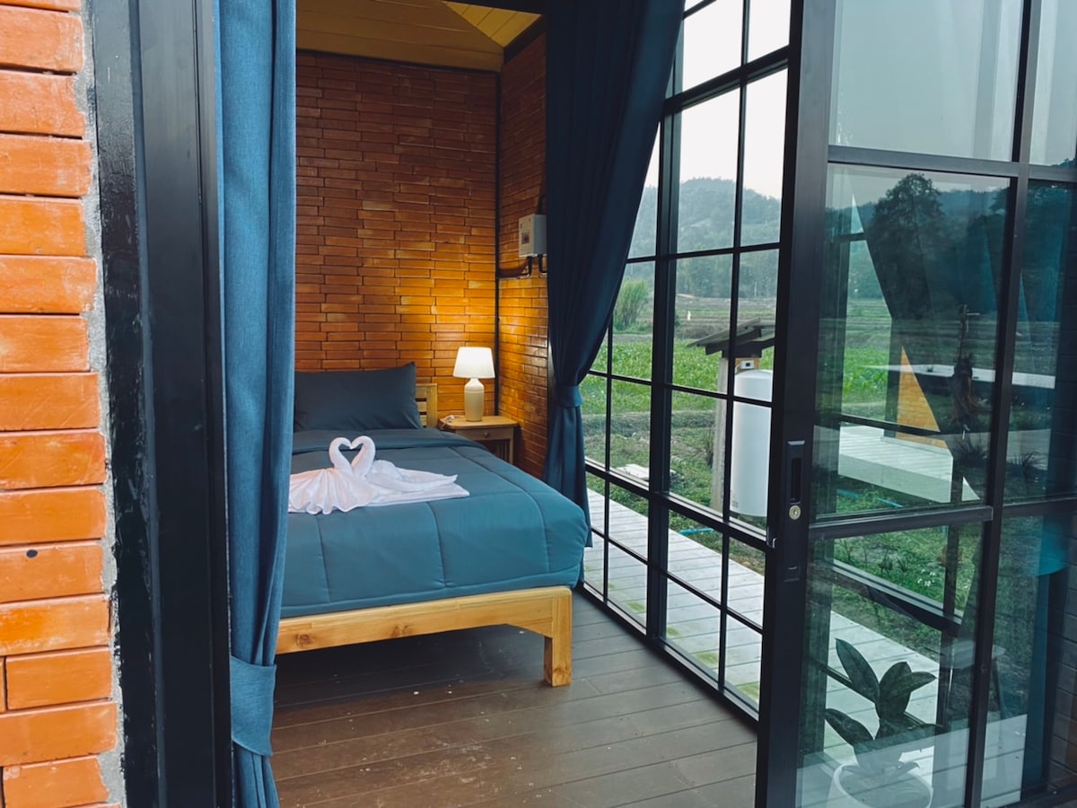 A cozy accommodation features a double bed with a soft blue blanket. A bedside lamp provides gentle illumination. Large glass doors lead to a view of lush green fields and mountains, framed by dark blue curtains that add a touch of elegance.
