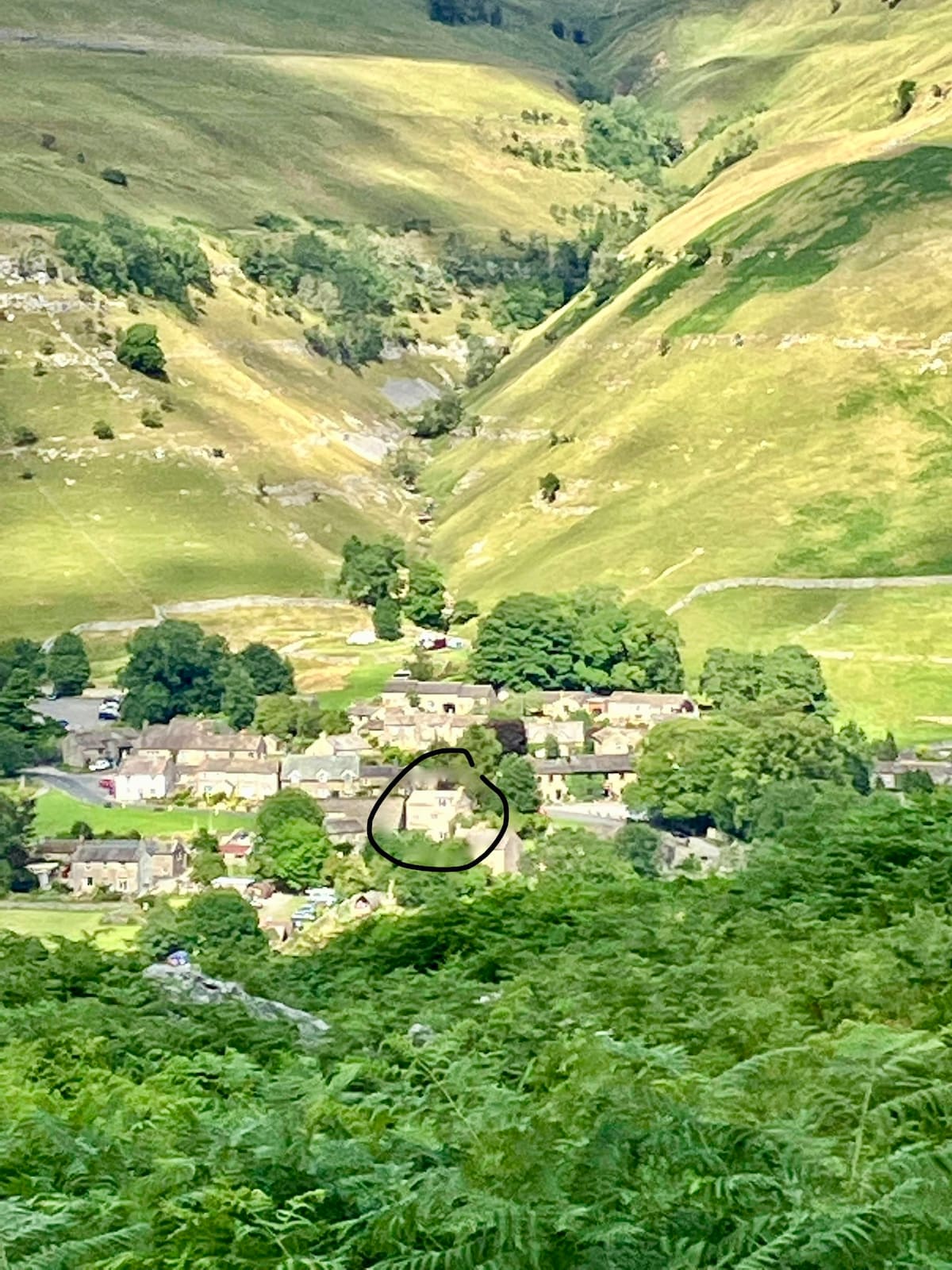 A panorama showcases the village nestled within a verdant valley, surrounded by rolling hills. The buildings, with stone facades, are interspersed with lush trees. The sunlight illuminates the landscape, highlighting the natural beauty of the Yorkshire Dales.