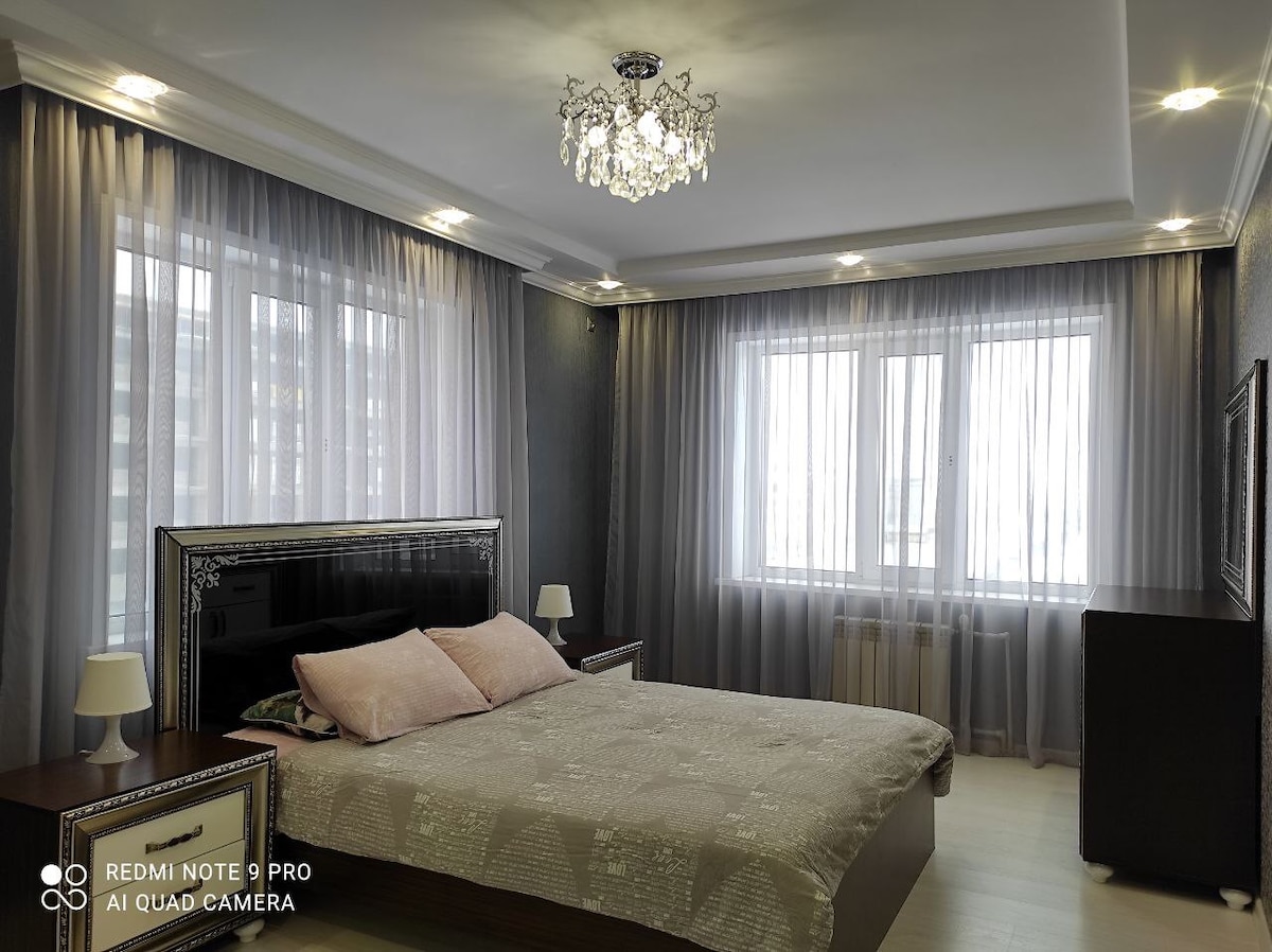 A spacious bedroom is illuminated by natural light streaming through large windows covered with sheer curtains. A comfortable bed is positioned at the center, flanked by bedside lamps. A stylish chandelier hangs from the ceiling, adding an elegant touch to the ambiance.