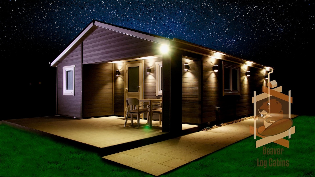 The modern log cabin is illuminated by exterior lights, showcasing a spacious patio with a table and chairs. The surrounding area is grassy, while a starry sky serves as a backdrop, emphasizing the cabin's tranquil setting.